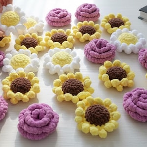 May include: A collection of crocheted flowers in various colours and shapes, including yellow and brown sunflowers, white and yellow daisies, and solid purple rose-like blossoms, all arranged on a flat surface for display and crafting purposes.