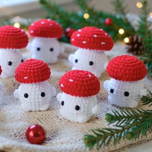 May include: A collection of small, crocheted mushroom figures, each featuring a bright red cap with white stitched spots and a white stem. The figures have black bead eyes and subtle pink stitched cheeks, arranged in a group on a textured surface.