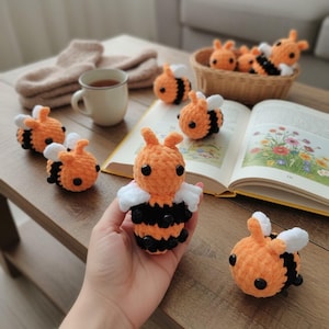 May include: Several small, handmade crochet bees featuring orange and black yarn bodies with white yarn wings and black bead eyes. One bee is held in the foreground, while others are scattered on a wooden surface near a book and a woven basket.