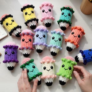 Puede incluir: Una colección de doce juguetes de peluche con forma de lápiz hechos de hilo suave. Cada juguete presenta una cara sonriente con ojos de cuentas negras, una goma rosa, una sección central blanca y una punta negra, decorado con pequeñas cuentas.