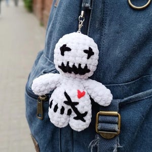 May include: A white crochet doll with black cross-shaped eyes and mouth, a red heart, and black stitched details. The doll is attached to a silver keychain and hangs from a blue denim backpack.