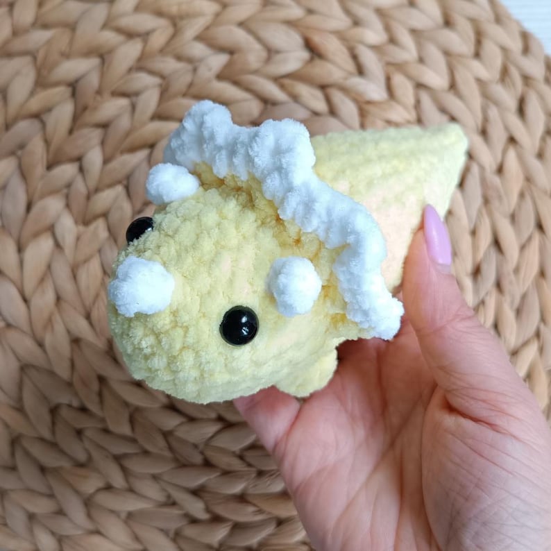 May include: A small, plush triceratops toy made of soft, yellow yarn with a white, textured frill and horns. The dinosaur features two round, black plastic eyes and a rounded snout, crafted with a fuzzy, tactile crochet texture throughout.