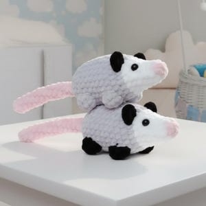 May include: Two handmade crochet opossums. The opossums are grey with black ears and feet, white faces, and pink noses and tails. One opossum is stacked on top of the other. The toys are on a white surface.