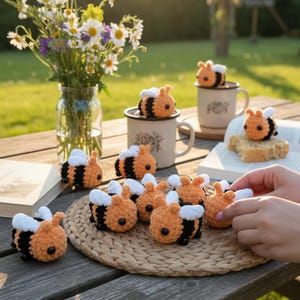 Könnte beinhalten: Eine Sammlung kleiner, handgefertigter Häkelbienen mit orange-schwarz gestreiften Körpern, weißen Flügeln und schwarzen Perlenaugen. Mehrere Bienen sind auf einer runden geflochtenen Matte angeordnet, weitere sitzen bei Tassen und Büchern.