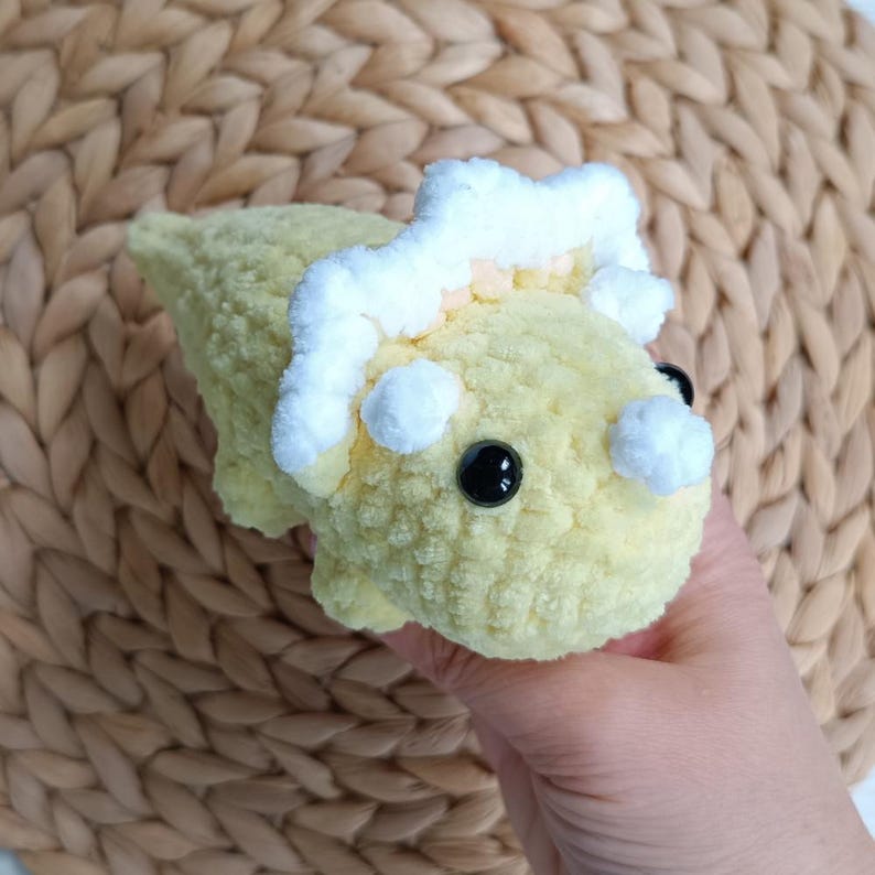 May include: A small, handmade crochet dinosaur toy crafted from soft, pale yellow yarn. The creature features a prominent white, textured frill around its head, a small white horn on its snout, and two round, glossy black plastic safety eyes.