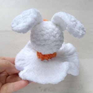 Puede incluir: Un peque&ntilde;o juguete de peluche tejido a ganchillo en color blanco, con dos orejas largas y ca&iacute;das, una cabeza redonda y una base ancha de falda blanca con volantes. Se ve una peque&ntilde;a secci&oacute;n de hilo naranja brillante en el centro de la uni&oacute;n.
