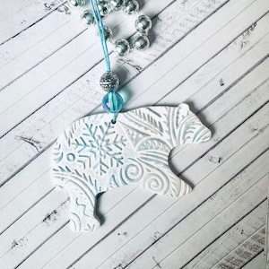 May include: White ceramic bear ornament with a blue and white snowflake and floral design. The ornament is hanging from a silver beaded necklace with a blue bead.