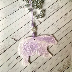 May include: A purple and white ceramic bear ornament with a snowflake design. The ornament is hanging from a purple cord with white beads.