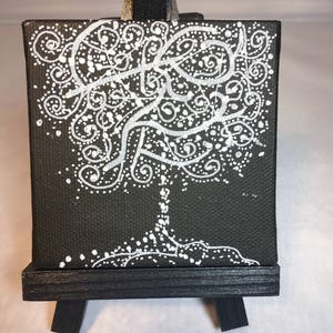 May include: A small black canvas painting with a white tree design. The tree is made up of swirls and dots. The painting is on a small black easel.