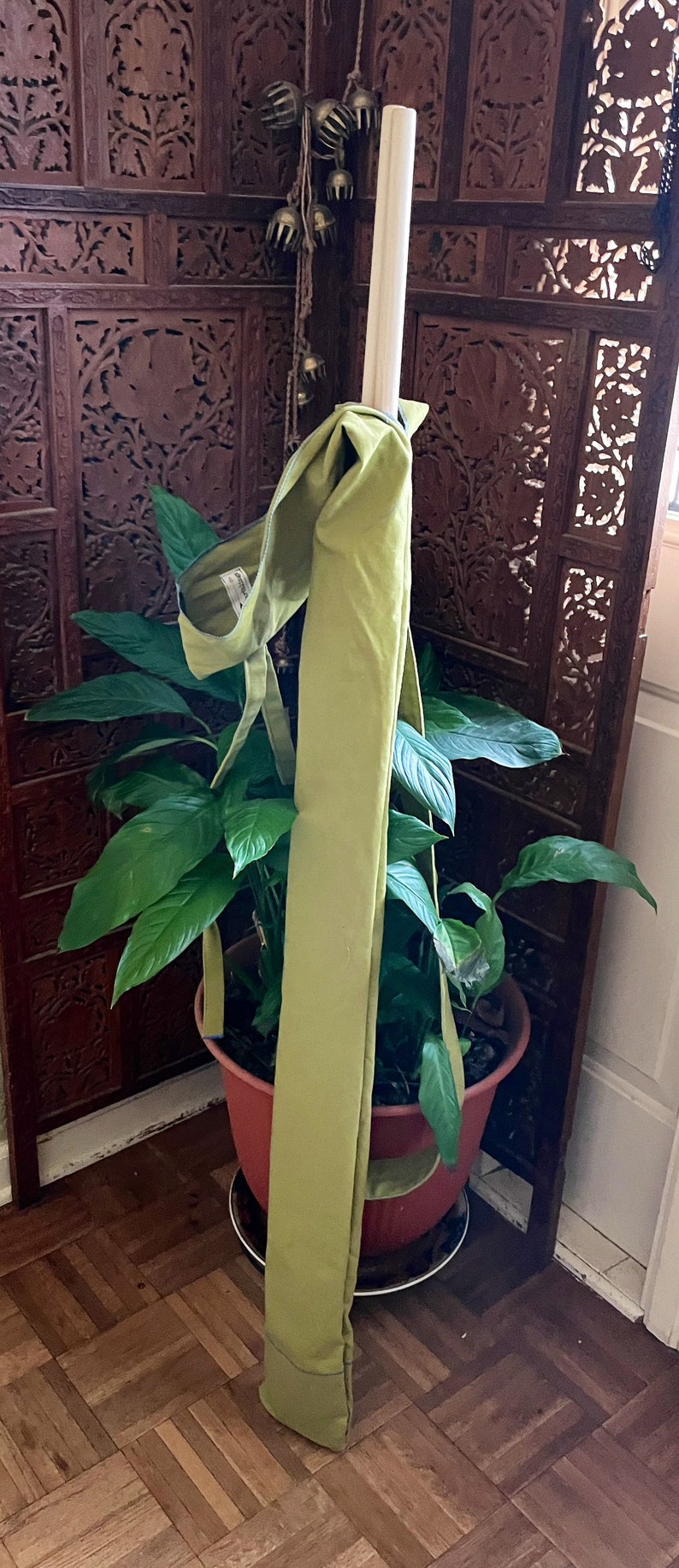 May include: A green fabric yoga mat rolled up and tied with a brown strap, leaning against a wooden screen with intricate carvings. The mat is sitting in a terracotta pot with a green plant.