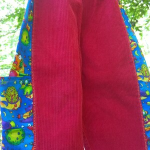 May include: Red corduroy pants with a blue and yellow patterned fabric trim. The trim features a cartoon design of space creatures and planets.