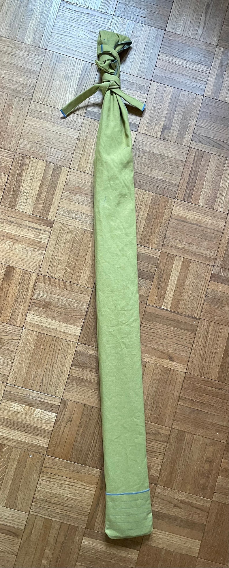 May include: A long, green fabric bag tied with a blue ribbon. The bag is lying flat on a wooden floor.