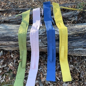 May include: Five fabric belts in different colors, including green, pink, blue, and yellow, are laid out on a log. The belts are all made of a similar fabric and have a simple design.