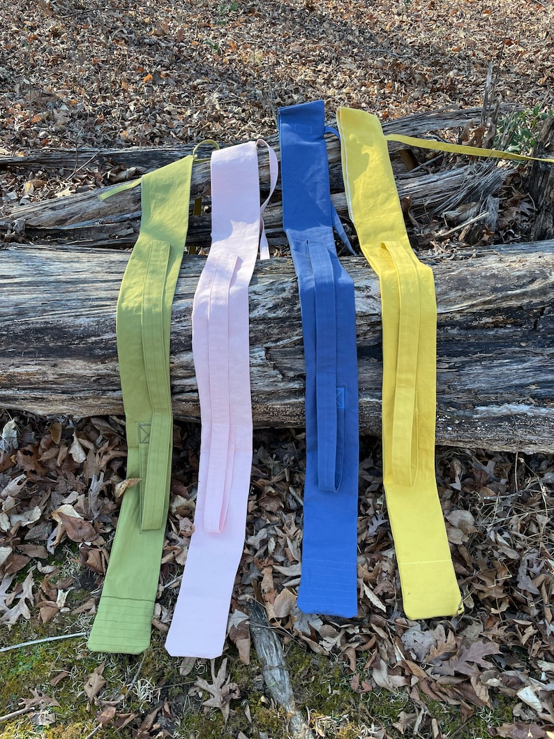 May include: Five fabric belts in different colors, including green, pink, blue, and yellow, are laid out on a log. The belts are all made of a similar fabric and have a simple design.