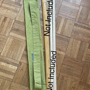May include: A green canvas bag with a blue trim and a long strap. The bag is lying on a wooden floor with two wooden sticks next to it. The sticks are labeled "Not included".