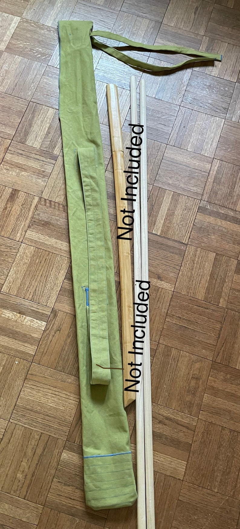 May include: A green canvas bag with a blue trim and a long strap. The bag is lying on a wooden floor with two wooden sticks next to it. The sticks are labeled "Not included".