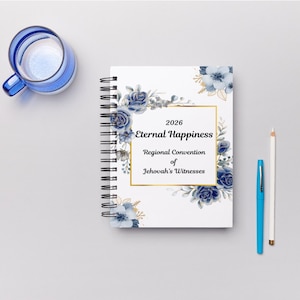 Puede incluir: Un cuaderno de espiral blanco con diseños florales azules y detalles dorados. La portada incluye el texto "2026 Eternal Happiness Regional Convention of Jehovah's Witnesses". También hay una taza azul, un bolígrafo azul y un lápiz blanco.