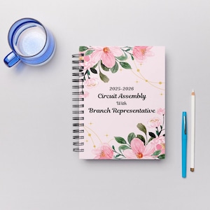 Könnte beinhalten: Ein spiralgebundenes Notizbuch mit hellrosa Einband mit floralen Designs und dem Text "2025-2026 Circuit Assembly With Branch Representative". Ein blauer Stift, ein weißer Bleistift und ein blau-weißes Glas sind ebenfalls sichtbar.
