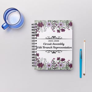 May include: A floral notebook with a spiral binding, featuring purple and lavender flowers and green leaves. The notebook has the text "2025-2026 Circuit Assembly With Branch Representative". A blue pen, white pencil, and a blue glass are also in the image.