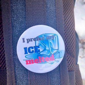 May include: A white button with the text "I prefer my ICE melted" in blue and red. The button features a blue graphic of an ice cube melting. The button is attached to a brown backpack.