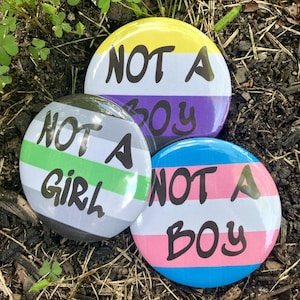Peut inclure: Trois badges ronds avec les phrases "NOT A BOY" et "NOT A GIRL" en texte noir. Les badges présentent les couleurs des drapeaux de la fierté non binaire, asexuelle et transgenre. Les badges sont posés sur un fond de paillis et de plantes vertes.