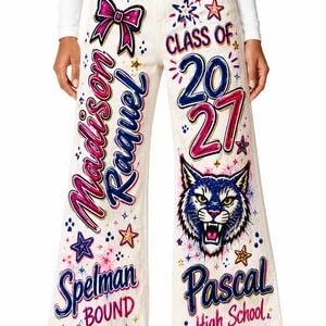 May include: Off-white wide-leg jeans featuring colorful airbrushed designs including a bow, stars, and the text Madison Raquel, Spelman Bound, Class of 2027, a wildcat face, and Pascal High School. The denim pants have a button closure at the waist.