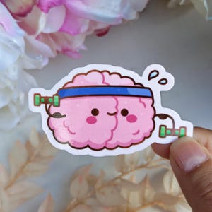 May include: A die-cut sticker featuring a cartoon brain character with a light pink color, wearing a blue headband, and lifting small green dumbbells. The brain has a smiling face with rosy cheeks and two small sweat droplets above it.