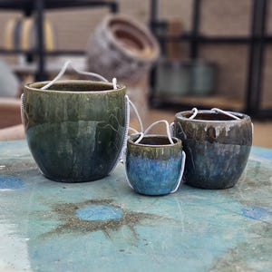 May include: Three ceramic hanging planters in varying sizes. The planters have a glossy finish with shades of green and blue. Each planter is suspended by a white rope. The planters are displayed on a light blue surface.