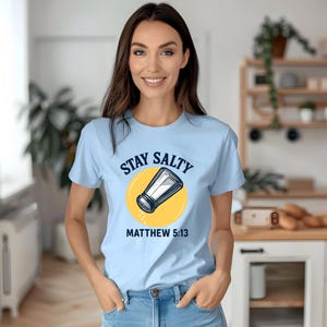 Può includere: Una maglietta azzurra con la scritta "STAY SALTY" sopra un'illustrazione di un saliera su un cerchio giallo. Il testo "MATTEO 5:13" è sotto. La maglietta è indossata da una persona.