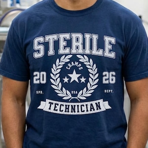 May include: A navy blue short-sleeve t-shirt featuring a white graphic print. The design includes the text STERILE TECHNICIAN, a laurel wreath surrounding a central star, the numbers 20 and 26, and the abbreviations SPD and DEPT. USA is printed.