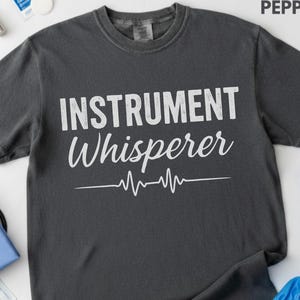 May include: A charcoal gray short-sleeve t-shirt featuring the white text "INSTRUMENT Whisperer" printed across the chest, accompanied by a white EKG heartbeat line graphic centered below the text. The shirt has a crew neckline and a standard fit.