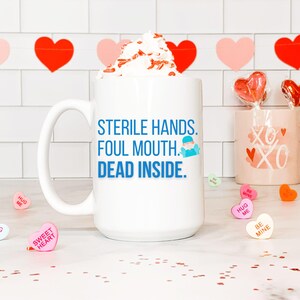 May include: A white ceramic mug with the text "STERILE HANDS. FOUL MOUTH. DEAD INSIDE." in blue. The mug is filled with whipped cream and sprinkles. Valentine's Day candy hearts and a pink mug with "XO" are also visible.