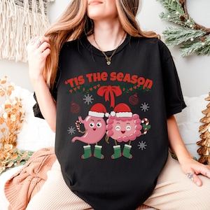 Op de afbeelding: Zwart T-shirt met de tekst "'Tis the Season" in rood en groen. Het shirt heeft cartoon illustraties van een maag en darmen met kerstmutsen en zuurstokken, met sneeuwvlokken en ornamenten.