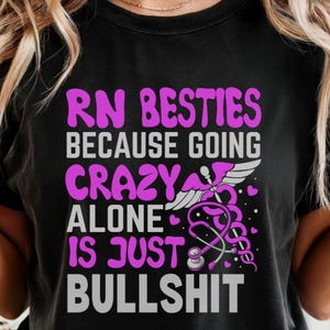 以下が含まれることがあります： 黒いTシャツ。テキスト「RN Besties Because Going Crazy Alone Is Just Bullshit」が白とピンクでプリントされています。聴診器とカドゥケウスのグラフィックも特徴です。