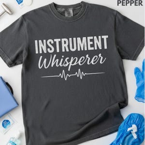 Może przedstawiać: Szara koszulka w kolorze pieprzu z białym napisem "INSTRUMENT Whisperer" i białą grafiką linii przypominającą bicie serca. Koszulka ma okrągły dekolt i wydaje się być wykonana z miękkiego materiału.