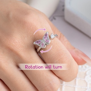 May include: A silver-colored ring featuring a butterfly design with pink accents and a small, clear gemstone. The ring has a rotating feature, as indicated by the text "Rotation will turn."