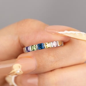 May include: A silver ring with a line of square-cut gemstones in rainbow colours. The gemstones are arranged in a spectrum, including purple, green, blue, yellow, pink, and orange. The ring is held against a neutral grey background.