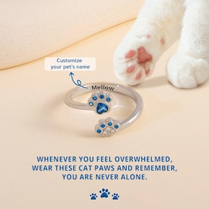 May include: A silver-colored adjustable ring with two paw-shaped designs, each adorned with blue and clear gemstones. The ring is engraved with the name "Mellow". The image includes the text "Customize your pet's name" and "Whenever you feel overwhelmed, wear these cat paws and remember, you are never alone."