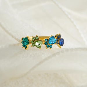 May include: A gold-toned ring featuring a series of colorful gemstones. The ring is adorned with teal, green, light blue, and dark blue stones, arranged in a decorative pattern. The gemstones are cut in various shapes, adding to the ring's visual appeal.
