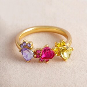 May include: A gold-colored ring featuring three heart-shaped gemstones in purple, pink, and yellow. The ring has a delicate design with smaller accent stones surrounding each heart, creating a colorful and eye-catching piece of jewelry.