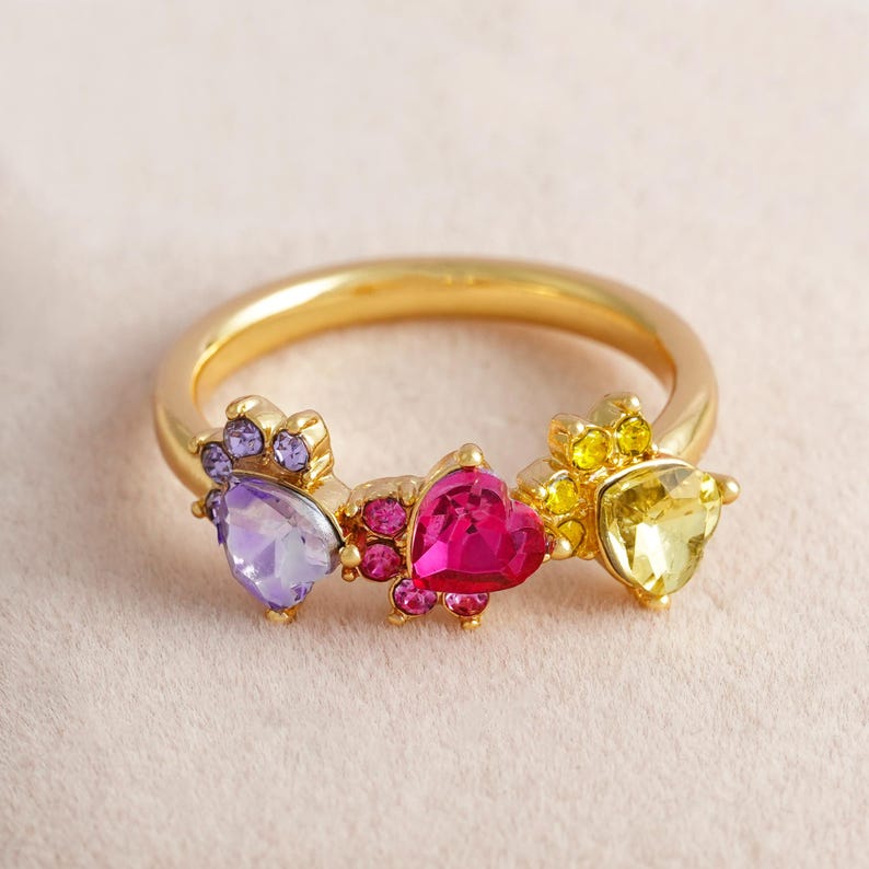 May include: A gold-colored ring featuring three heart-shaped gemstones in purple, pink, and yellow. The ring has a delicate design with smaller accent stones surrounding each heart, creating a colorful and eye-catching piece of jewelry.