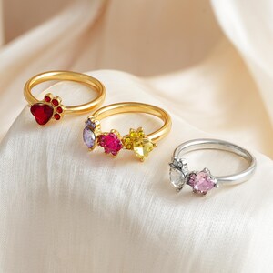 May include: Three rings with gemstone accents. One gold ring features a red heart-shaped stone with a paw print design. Another gold ring has purple, pink, and yellow stones. A silver ring has clear and pink heart-shaped stones.