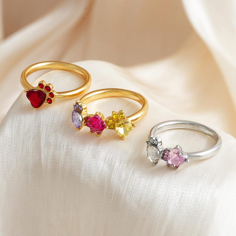 May include: Three rings with gemstone accents. One gold ring features a red heart-shaped stone with a paw print design. Another gold ring has purple, pink, and yellow stones. A silver ring has clear and pink heart-shaped stones.