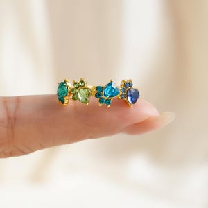 May include: A gold-toned ring featuring a series of colorful gemstones in shades of teal, green, and blue. The gemstones are arranged in a decorative pattern, creating a unique and eye-catching design. The ring is held by a finger.