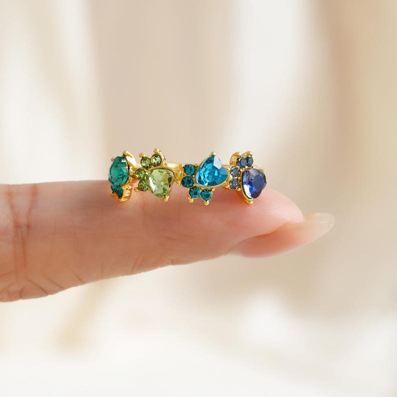 May include: A gold-toned ring featuring a series of colorful gemstones in shades of teal, green, and blue. The gemstones are arranged in a decorative pattern, creating a unique and eye-catching design. The ring is held by a finger.