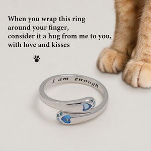 May include: A silver-colored ring with a paw-shaped design and blue gemstones. The ring is engraved with the words "I am enough." The image also includes text that reads "When you wrap this ring around your finger, consider it a hug from me to you, with love and kisses."