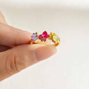 May include: A gold-colored ring featuring three gemstone settings. The gemstones are purple, pink heart-shaped, and yellow, set in a floral design. The ring is held by a person's fingers.