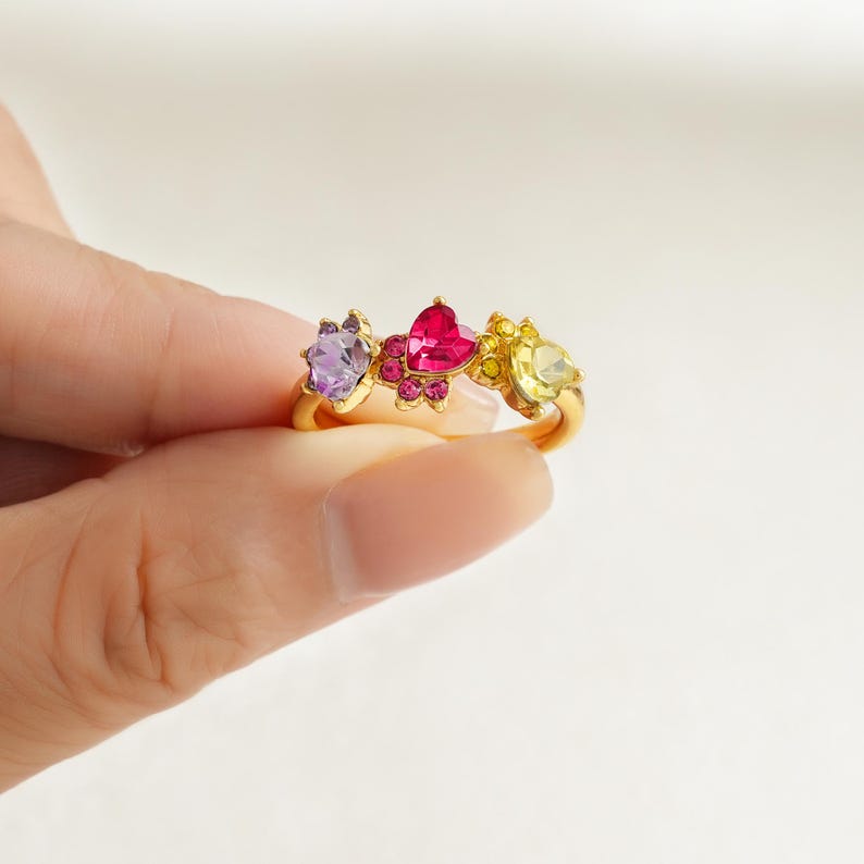May include: A gold-colored ring featuring three gemstone settings. The gemstones are purple, pink heart-shaped, and yellow, set in a floral design. The ring is held by a person's fingers.