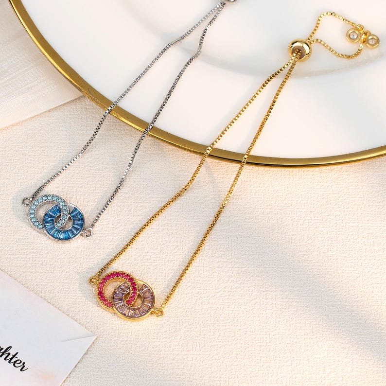May include: Two delicate bracelets with interlocking circles. One bracelet is silver with blue crystals, the other is gold with pink and clear crystals. The bracelets are displayed on a white plate with a gold rim, showcasing their elegant design.