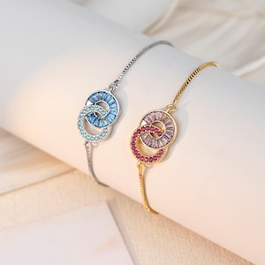 May include: Two bracelets with interlocking circles, one silver with blue stones, the other gold with pink and red stones. The bracelets are displayed on a white surface, showcasing the jewelry's design and color variations.